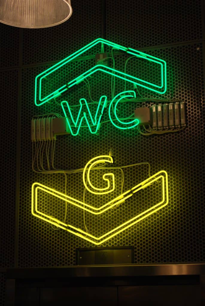 Large neon sign placed over the lift. at the top is the outline of a green arrow head, pointing upwards with the word WC underneath. Below this is yellow is the letter G followed by a downward pointing arrow head
