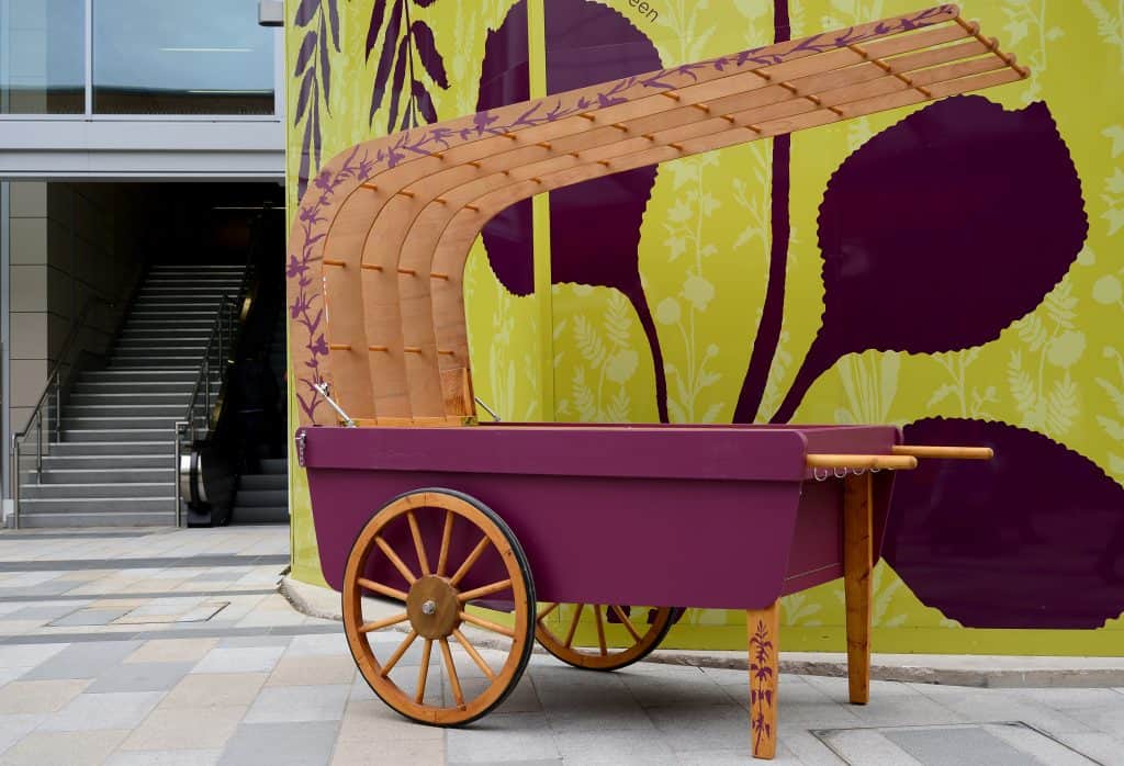 Community barrow, based on a traditional costermonger barrow with wheels and wooden overarching canopy. The base is painted in aubergine and the rest is left in natural wood with a painted leaf motif in aubergine snaking up the leg rests and the canopy edge.