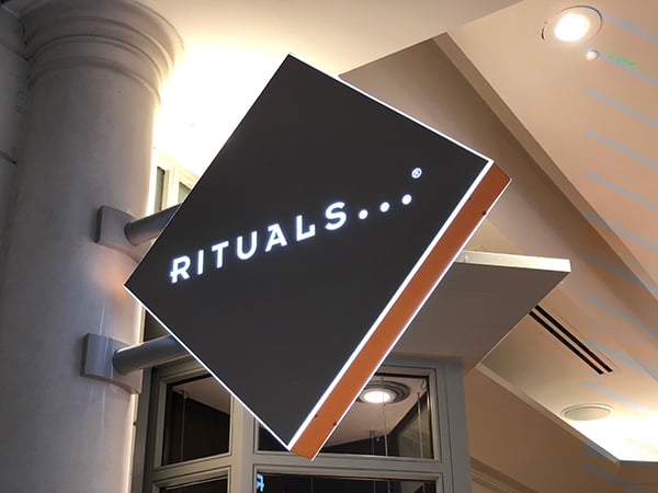 Photo of a diamond shaped retailer branding sign projecting from a column.The sign is faced in grey with white lettering and copper edges