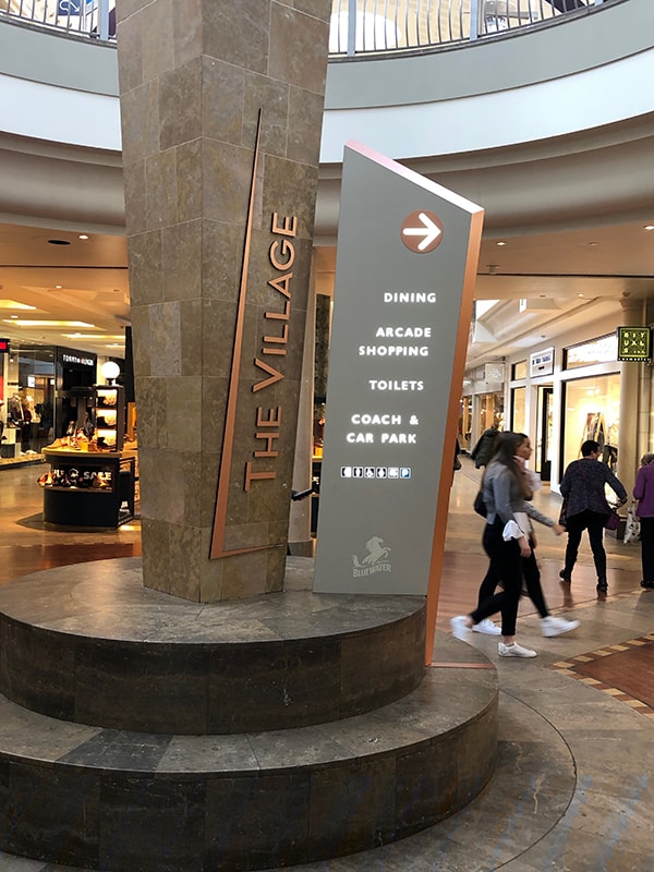 Large totem sign at the entrance to The Village Arcade. The sign is faced in a grey beige colour with a coppery coloured chamfered sides. The content is in white with a copper coloured disc surrounding the directional arrow.