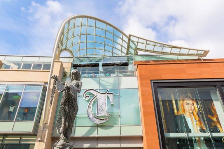 Large letter T in a gothic style, applied above the entrance to The Trinity Leeds shopping centre. The sign is formed from a stainless steel outline and filled with individual metal discs to give it sparkle. In front of the sign is a metal mesh sculpture of the goddess Minerva