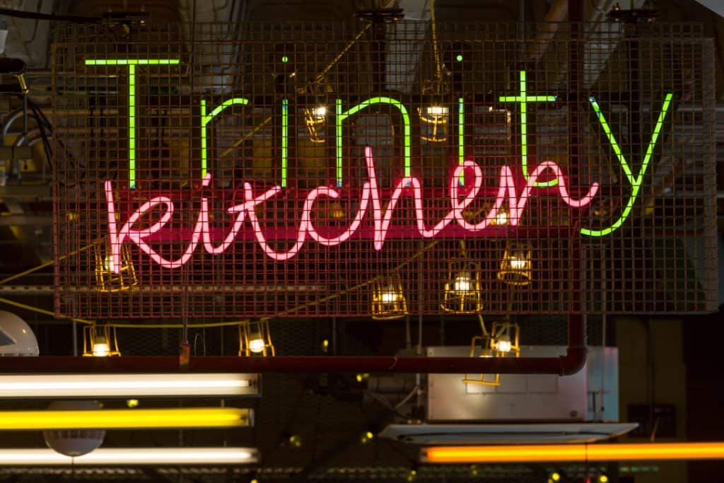 Close up of the Trinity Kitchen identification sign featuring green illuminated individual letters spelling out Trinity, overlaid by the word 'kitchen' in pink neon script. There is a metal chicken-wire sheet in front of the sign and a string of small industrial inspection lights behind. The fixings for the signs and infrastructure for the services is clearly visible.