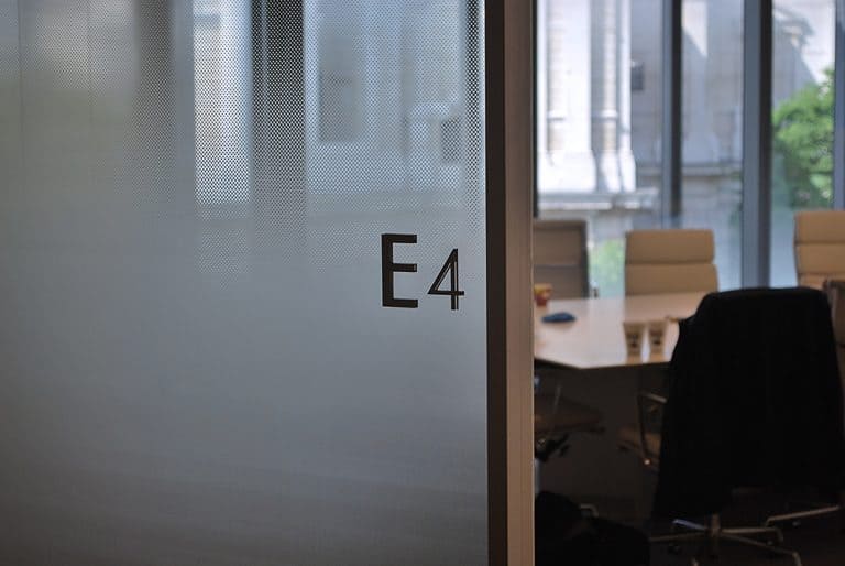 View through the open door to an office. The glazed office wall is covered in manifestation which has a diffused pattern design. The room name E4 is applied to the glass as individual characters