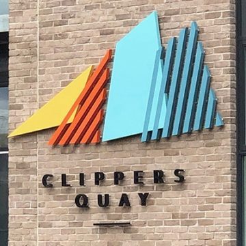 Multicoloured identity sign for Clippers Quay applied to a brick wall. The letters are black and applied as individual 3D characters. The logo type is composed of four overlapping sails – a yellow solid triangle, followed by an orange triangular shaped striated outline, a solid pale blue trapezoid and a further turquoise striated trapezoid.